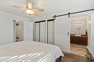 Tiled bedroom featuring crown molding, ceiling fan, connected bathroom, a barn door, and multiple closets