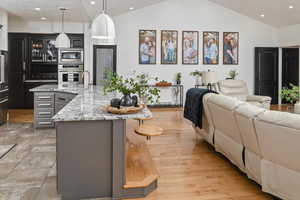 Kitchen with vaulted ceiling, stainless steel microwave, an island with sink, hanging light fixtures, and light stone counters