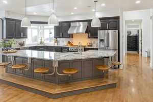 Kitchen with pendant lighting, wall chimney range hood, a breakfast bar area, high quality fridge, and an island with sink