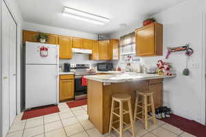 Kitchen with stainless steel electric stove, a breakfast bar area, white refrigerator, light tile patterned floors, and kitchen peninsula