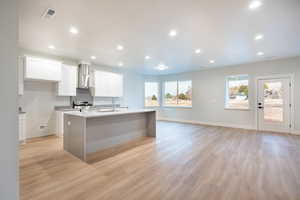 Kitchen with white cabinetry featuring light waterproof luxury vinyl plank flooring, stainless steel range, a kitchen island made of elegant quartz waterfall counter tops with sink, and wall chimney range hood