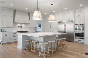 Kitchen with an island with sink, sink, white cabinets, custom exhaust hood, and built in appliances