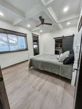 Bedroom featuring coffered ceiling, light wood-type flooring, beamed ceiling, ceiling fan, and a barn door