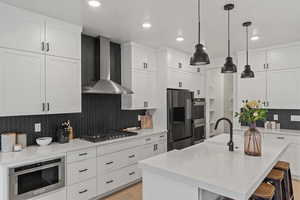 Kitchen featuring appliances with stainless steel finishes, an island with sink, decorative backsplash, hanging light fixtures, and wall chimney exhaust hood