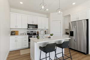 Kitchen with appliances with stainless steel finishes, white cabinetry, hanging light fixtures, light hardwood / wood-style floors, and a center island with sink