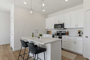 Kitchen with white cabinetry, hanging light fixtures, stainless steel appliances, an island with sink, and light wood-type flooring