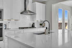 Kitchen with appliances with stainless steel finishes, sink, white cabinets, light stone counters, and wall chimney exhaust hood