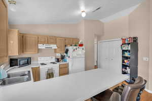 Kitchen with light brown cabinetry, lofted ceiling, sink, kitchen peninsula, and white appliances