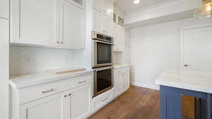 Kitchen with white cabinetry, decorative light fixtures, double oven, light stone countertops, and backsplash