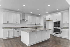 Kitchen with appliances with stainless steel finishes, white cabinetry, an island with sink, sink, and wall chimney range hood