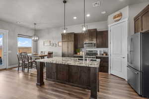 Kitchen with decorative light fixtures, dark brown cabinetry, stainless steel appliances, light stone countertops, and a center island with sink