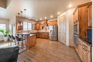 Kitchen featuring decorative light fixtures, a kitchen bar, stainless steel appliances, custom range hood, and light hardwood / wood-style flooring