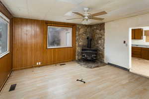 Unfurnished living room with ceiling fan, a wood stove, wooden walls, and light hardwood / wood-style floors