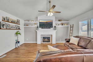 Living room with vaulted ceiling, built in features, a large fireplace, ceiling fan, and light hardwood / wood-style floors