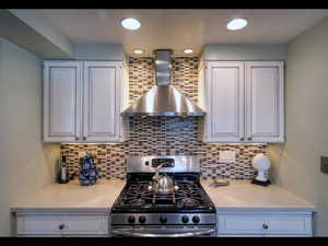 Kitchen featuring wall chimney exhaust hood, gas stove, decorative backsplash, and white cabinets