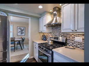 Kitchen featuring white cabinetry, ornamental molding, appliances with stainless steel finishes, and wall chimney range hood