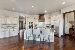 Kitchen with premium range hood, a kitchen island with sink, stainless steel appliances, light stone countertops, and cream cabinetry