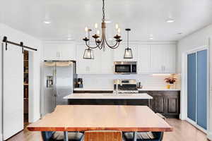 Kitchen with hanging light fixtures, a center island with sink, stainless steel appliances, a barn door, and white cabinets