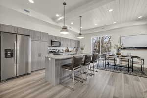 Kitchen featuring hanging light fixtures, a kitchen island with sink, light stone counters, stainless steel appliances, and light wood-type flooring