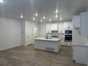 Kitchen featuring dark wood-type flooring, white cabinetry, stainless steel appliances, an island with sink, and wall chimney exhaust hood