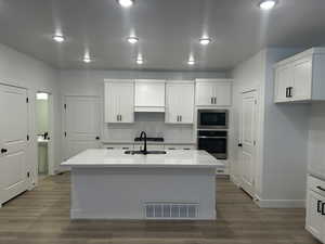 Kitchen featuring built in microwave, sink, white cabinetry, stainless steel oven, and an island with sink