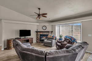 Living room with lofted ceiling, a stone fireplace, a textured ceiling, ceiling fan, and light hardwood / wood-style floors