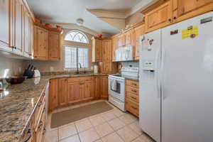 Kitchen featuring light tile patterned flooring, lofted ceiling, sink, white appliances, and dark stone counters