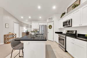 Kitchen featuring light tile patterned floors, appliances with stainless steel finishes, a kitchen island with sink, white cabinetry, and vaulted ceiling