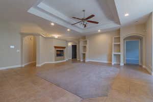 Unfurnished living room with ceiling fan, a tray ceiling, built in features, and light tile patterned floors