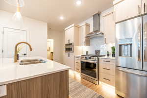 Kitchen with pendant lighting, sink, appliances with stainless steel finishes, wall chimney exhaust hood, and light wood-type flooring