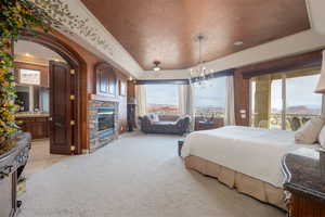 Carpeted bedroom featuring an inviting chandelier, connected bathroom, a stone fireplace, and a raised ceiling