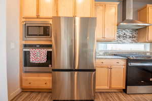 Kitchen with wall chimney range hood, light hardwood / wood-style flooring, backsplash, stainless steel appliances, and light brown cabinetry