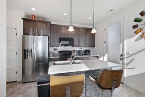 Kitchen with hanging light fixtures, a kitchen island with sink, dark brown cabinetry, black appliances, and light hardwood / wood-style flooring