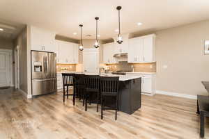 Kitchen with pendant lighting, stainless steel appliances, a kitchen island with sink, decorative backsplash, and white cabinets