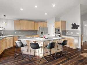 Kitchen with light brown cabinetry, decorative light fixtures, a center island, stainless steel appliances, and dark wood-type flooring