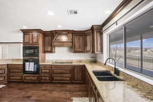 Kitchen with sink, dark hardwood / wood-style floors, light stone counters, black appliances, and custom range hood