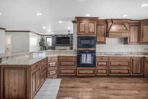 Kitchen with dark wood-type flooring, black appliances, crown molding, light stone countertops, and custom range hood