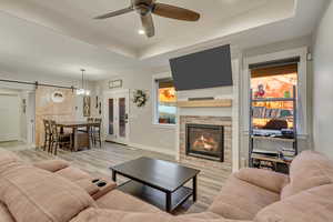 Living room with light hardwood / wood-style flooring, ceiling fan, a tray ceiling, a fireplace, and a barn door