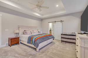 Bedroom with a barn door, ceiling fan, a tray ceiling, and light colored carpet