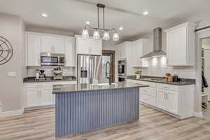 Kitchen featuring white cabinetry, hanging light fixtures, stainless steel appliances, built in microwave, double wall ovens, and wall chimney exhaust hood