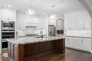 Kitchen featuring stainless steel appliances, a kitchen island with sink, white cabinets, and decorative light fixtures