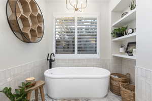 Bathroom with built in shelves, a bathing tub, tile walls, and a notable chandelier