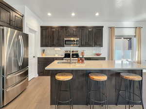 Kitchen featuring appliances with stainless steel finishes, backsplash, a kitchen bar, a kitchen island with sink, and light wood-type flooring