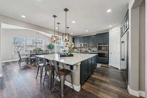 Kitchen with pendant lighting, appliances with stainless steel finishes, a kitchen island with sink, backsplash, and light stone counters