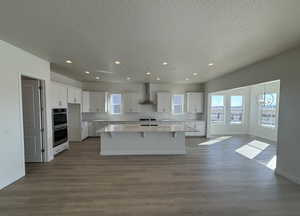 Kitchen with wall chimney range hood, white cabinetry, hardwood / wood-style floors, an island with sink, and stainless steel double oven