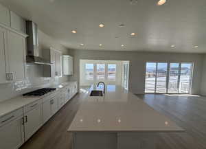 Kitchen with white cabinetry, wall chimney range hood, stainless steel gas stovetop, and a large island with sink