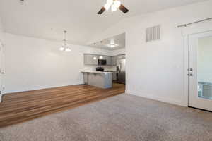 Unfurnished living room featuring medium toned carpet, ceiling fan, dining area with notable chandelier, and beautifully vaulted ceilings. Open floorplan to the Kitchen as well as access to patio door.