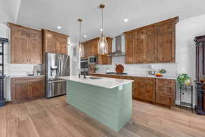 Kitchen with wall chimney range hood, sink, a kitchen island with sink, hanging light fixtures, and stainless steel appliances