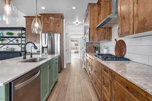 Kitchen with sink, hanging light fixtures, stainless steel appliances, light stone countertops, and wall chimney exhaust hood