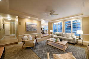 Living room with a raised ceiling, ornamental molding, ceiling fan with notable chandelier, and dark colored carpet